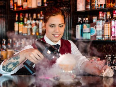 Skilled young female barkeeper using flavor bluster smoke gun while garnishing cocktail at bar counter