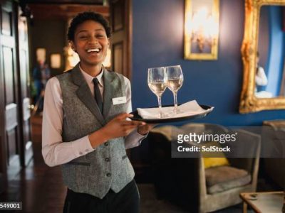 A well-dressed waitress is laughing and enjoying being at work, while she is holding wine glasses to be served to guests.
